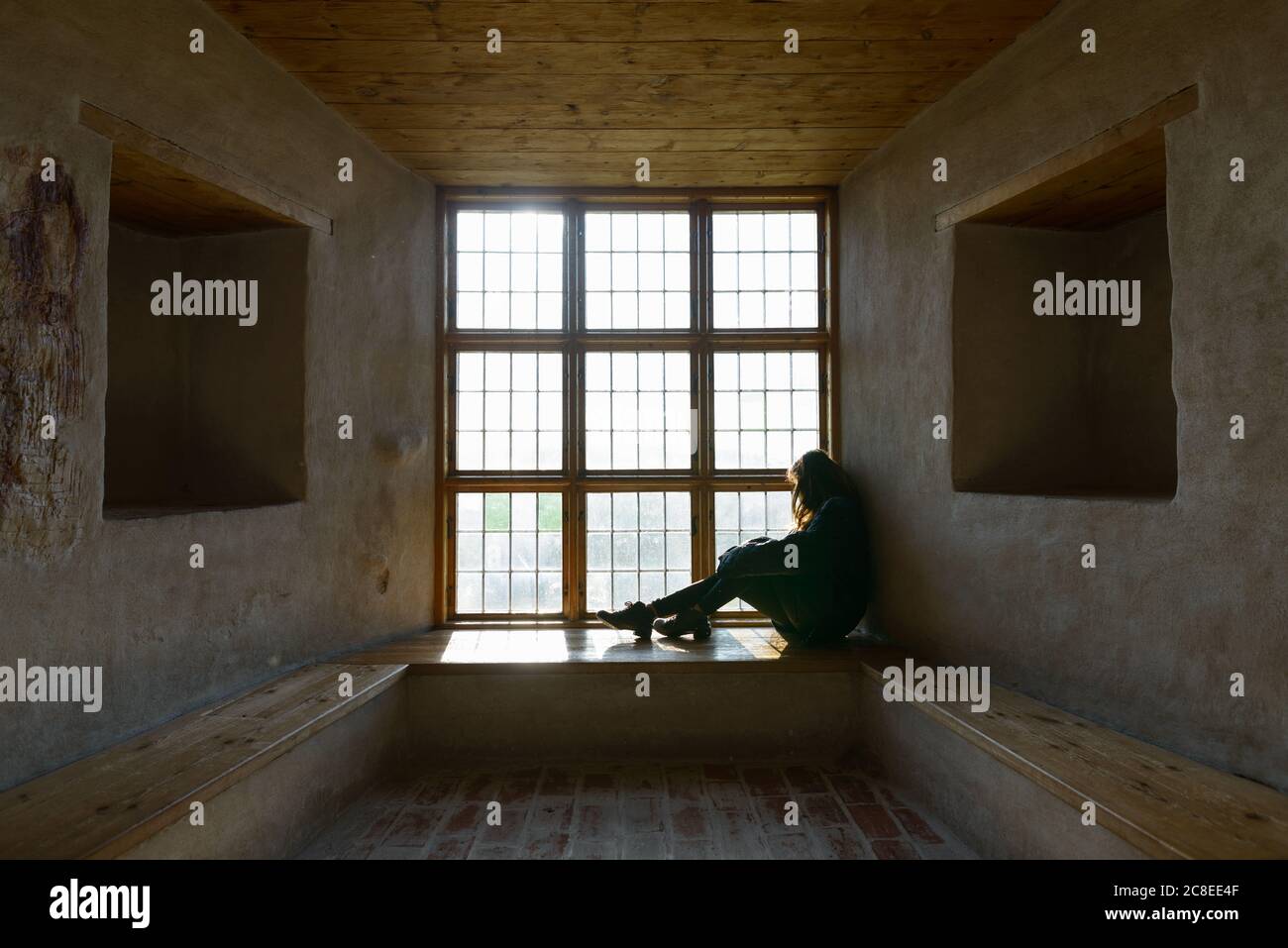 Junge Frau, die vor einem geschlossenen Holzfenster sitzt und das Sonnenlicht einströmt Stockfoto