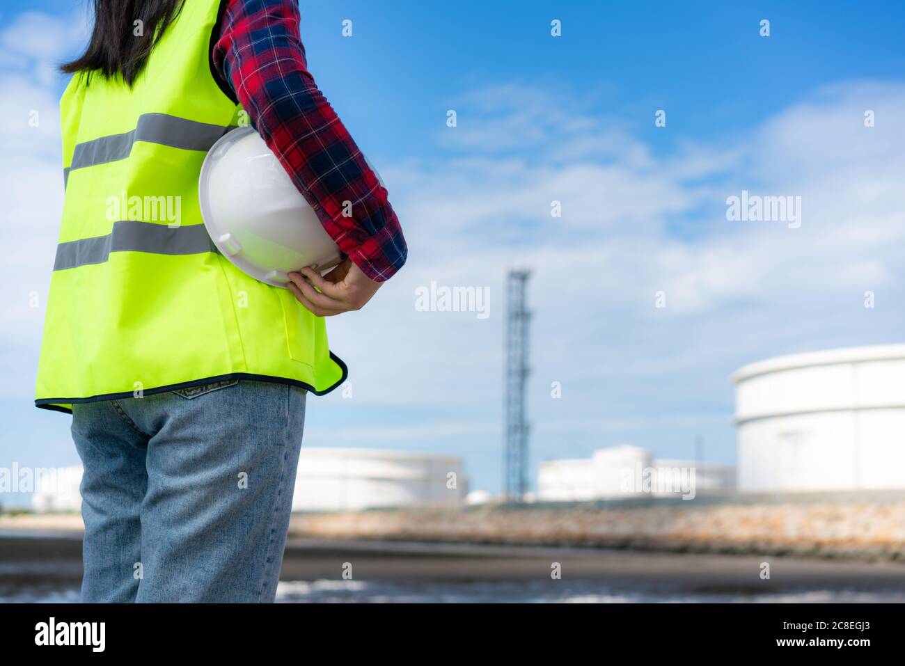 Asiatische Frau Ingenieur hält einen weißen Schutzhelm mit Öl Raffinerie Tanklager Fabrik im Hintergrund mit blauen Himmel. Stockfoto