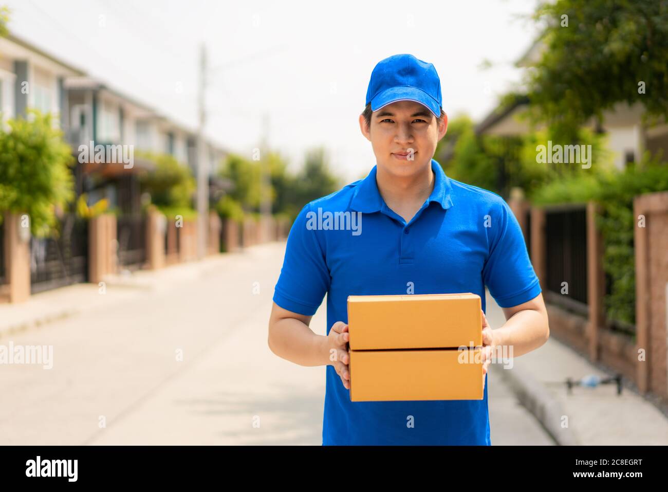 Asiatischer Auslieferungsjunge Mann in blauem, gleichmäßigem Lächeln und Kartonstapel im vorderen Hausdorf mit Kopfflächen. Werbung, Business, Tran Stockfoto