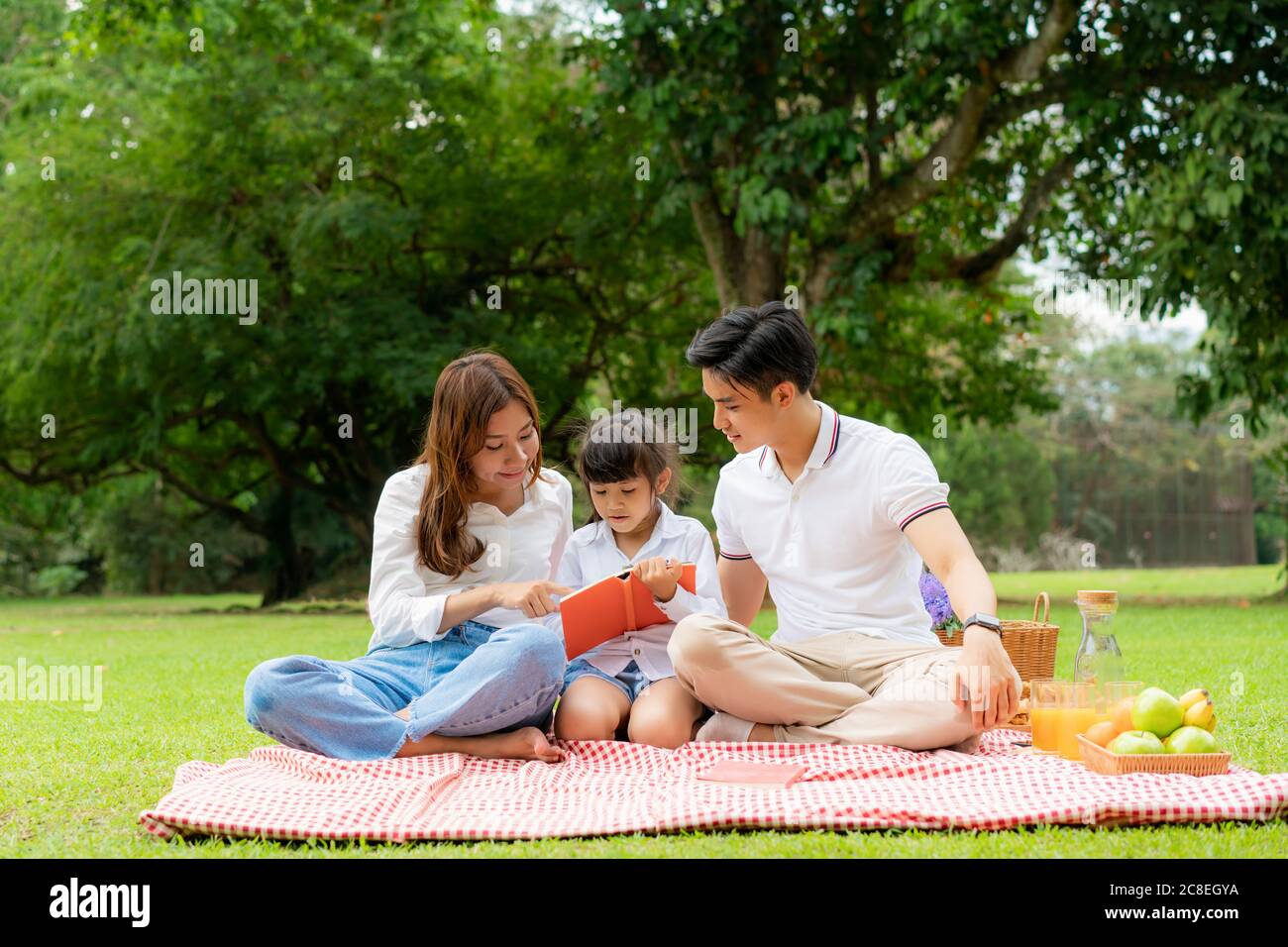Asiatische Teenager Familie glücklich Urlaub Picknick Moment im Park mit Vater, Mutter und Tochter Buch lesen zusammen im grünen Garten mit Obst und Essen. Stockfoto