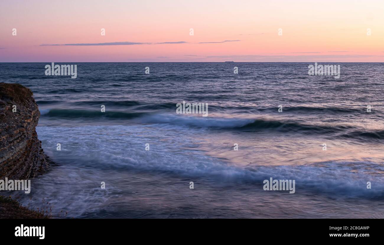 Ein Foto mit einer langen Verschlusszeit von einer Sekunde. Sanftes rosa Meer Sonnenuntergang. Glatte Wellen schlagen gegen die Felsen. Am Horizont sind Frachtschiffe zu sehen. Stockfoto