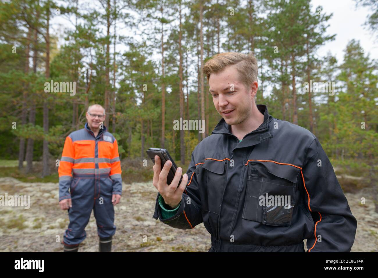 Glücklicher junger Mann hält Handy im Wald mit glücklichen reifen Mann im Hintergrund Stockfoto