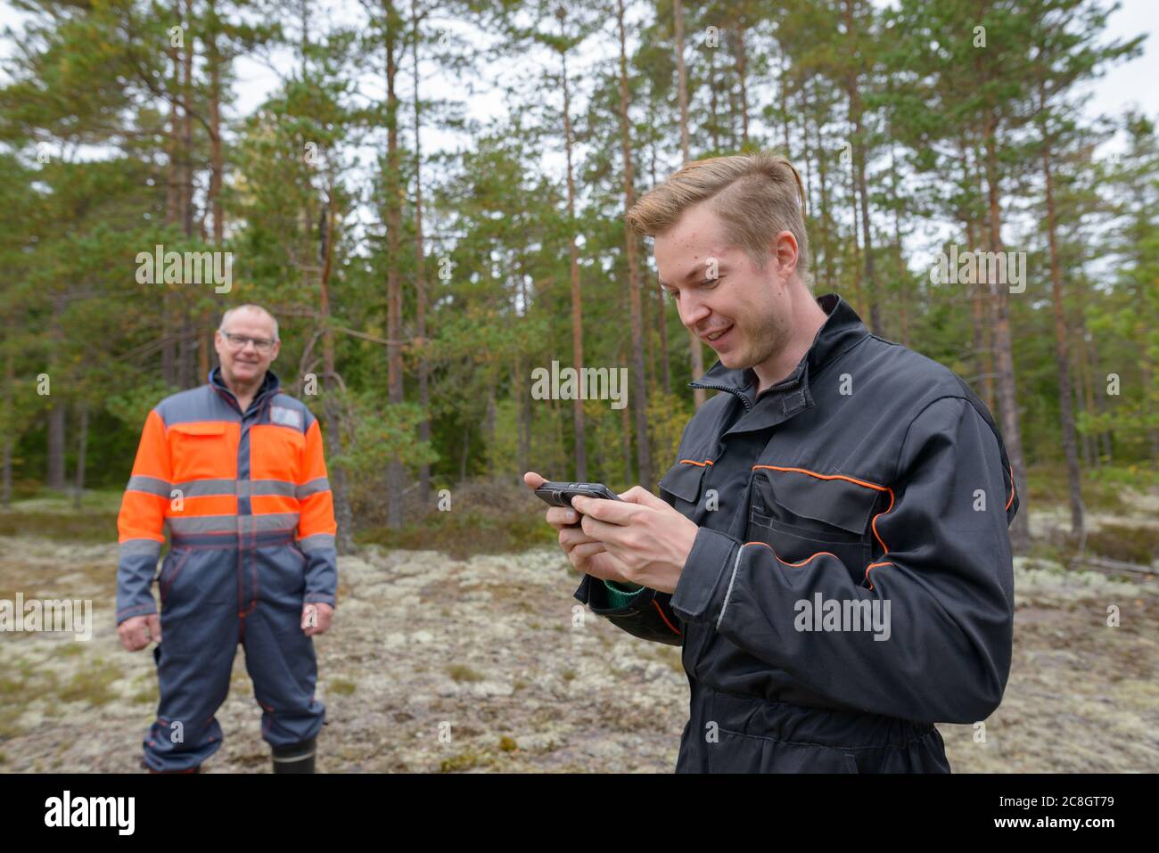 Glücklicher junger Mann mit Handy im Wald mit glücklichen reifen Mann im Hintergrund Stockfoto