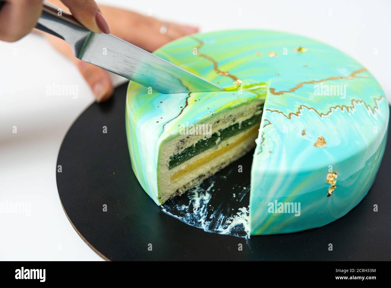 Nahaufnahme des Schneidens mit Messer Mousse Kuchen bedeckt blau und grün Spiegelglasur. Französisches Dessert. Gefrorener Spiegel Sahnehäubchen auf dem Kuchen. Backen und Konfektion Stockfoto