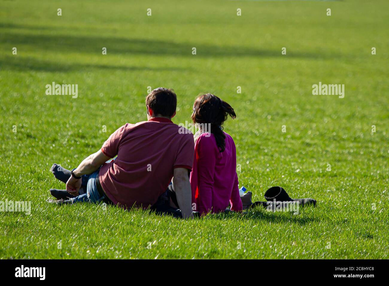 Ein junges Paar hat etwas Freizeit, während sie auf dem Gras sitzen und sich an einem sonnigen Nachmittag in einem Park entspannen. Mann trägt ein T-Shirt und Jeans, Frau wea Stockfoto