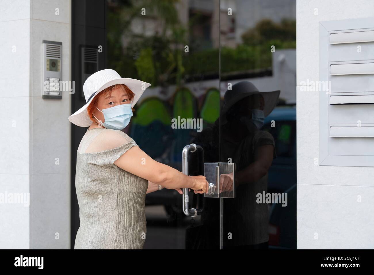 Reife asiatische Frau trägt eine chirurgische Maske und einen Hut greifen eine Tür Griff beim Blick auf die Kamera. Sicherheits- und Lifestyle-Konzept. Stockfoto
