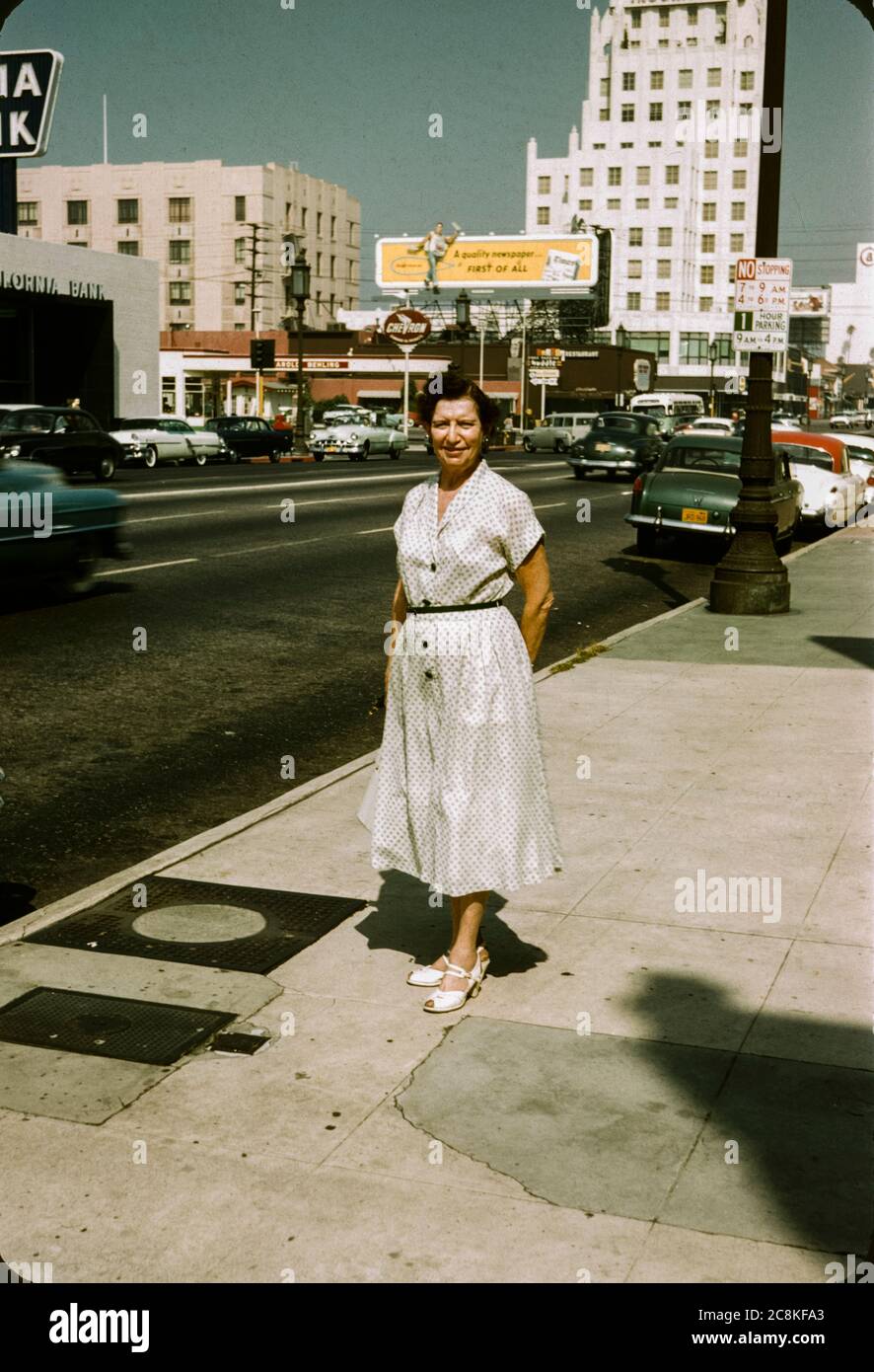 Frau, die in den 1950er Jahren auf dem Bürgersteig in der Innenstadt von Los Angeles, Kalifornien, stand. Plakate und 50er Jahre Autos sind im Hintergrund, einschließlich einer Plakatwand für die Los Angeles Times Zeitung. Stockfoto