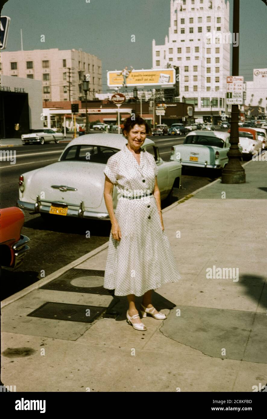 Frau, die 1957 auf dem Bürgersteig in der Innenstadt von Los Angeles, Kalifornien, stand. Plakate und 50er Jahre Autos sind im Hintergrund, einschließlich einer Plakatwand für die Los Angeles Times Zeitung. Los Angeles Street Scene 1957 Kodachrome Red Mount Cars Stockfoto