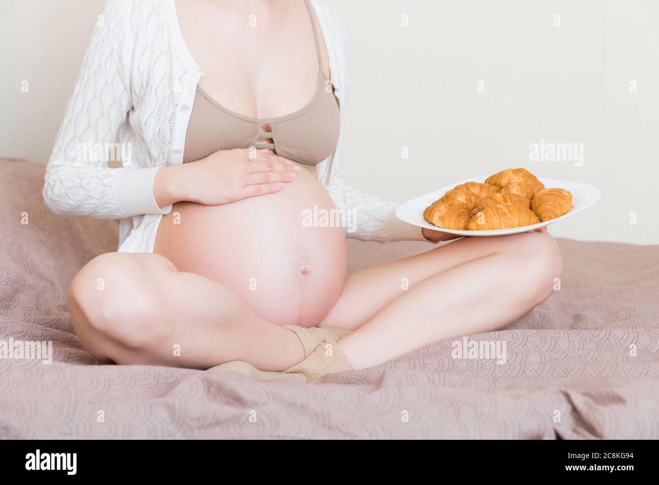 Nahaufnahme der jungen schwangeren Frau genießt essen Croissant im Bett. Ungesunde Gebäck während der Schwangerschaft Konzept. Stockfoto