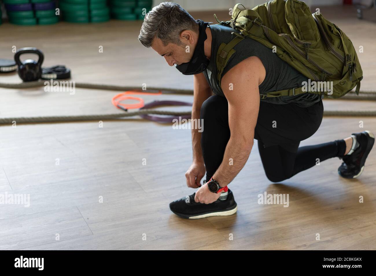 Motivierter Instruktor kniet sich nieder und bindet seine Schuhschnur mit Rucksack und Trainingsmaske. Beginn der Schulung. Für Boot Camp und Fitness-Konzept. Stockfoto