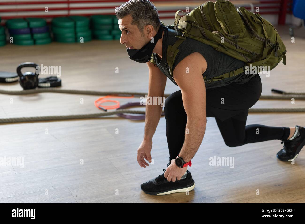 Motivierter Instruktor kniet sich nieder und bindet seine Schuhschnur mit Rucksack und Trainingsmaske. Beginn der Schulung. Für Boot Camp und Fitness-Konzept. Stockfoto