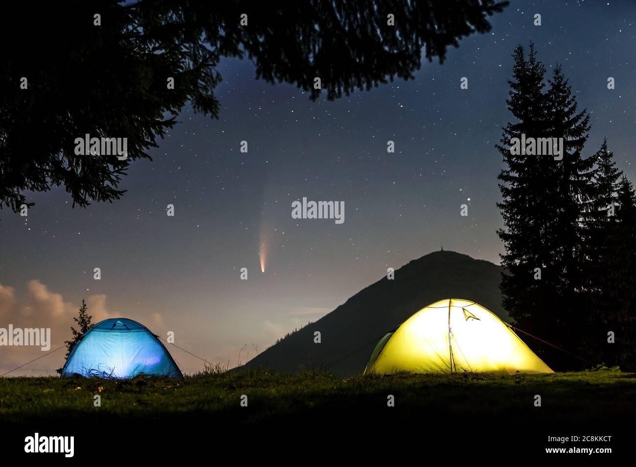 Zwei hell erleuchtete Touristenzelte auf Waldlichtung in Bergen mit Sternenhimmel und Neowise-Komet mit Lichtschweif. Stockfoto