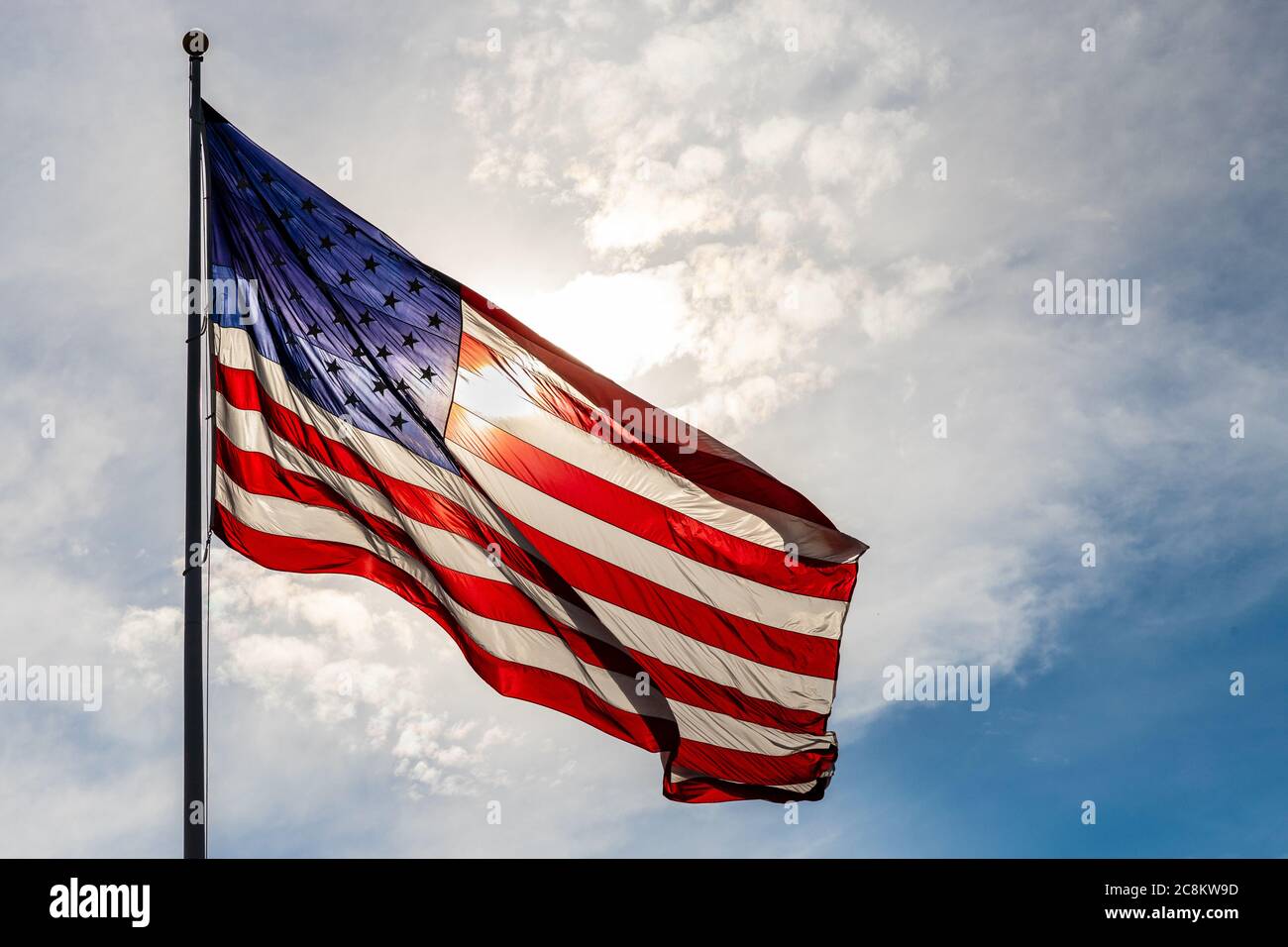 Große schöne amerikanische Flagge leuchtet von der Hintergrundbeleuchtung von der Sonne, gegen bewölkten Himmel Stockfoto