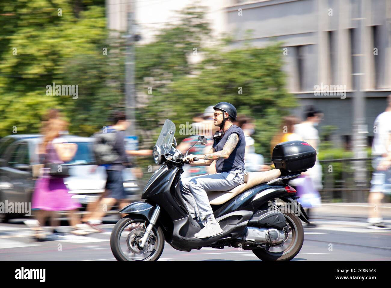 Belgrad, Serbien - 16. Juli 2020: Reifer tätowierte Mann, der im Stadtverkehr auf einem Motorroller mit dem Motorrad unterwegs ist, an der Kreuzung mit Menschen, die vorbei gehen Stockfoto