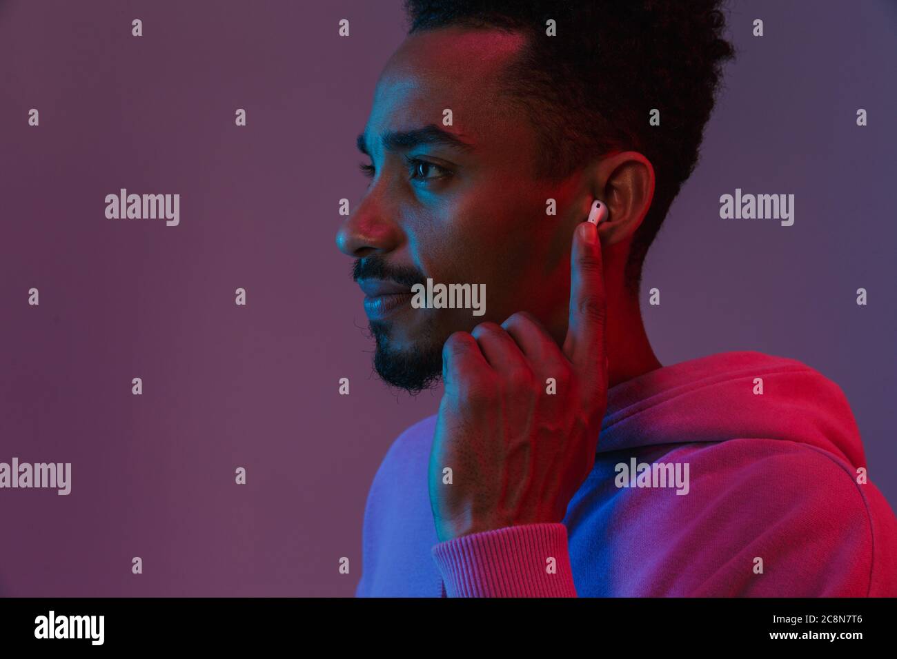 Portrait von Konzentriertem african american man in bunten hoodie Musik hören mit earpod über violett Hintergrund isoliert Stockfoto