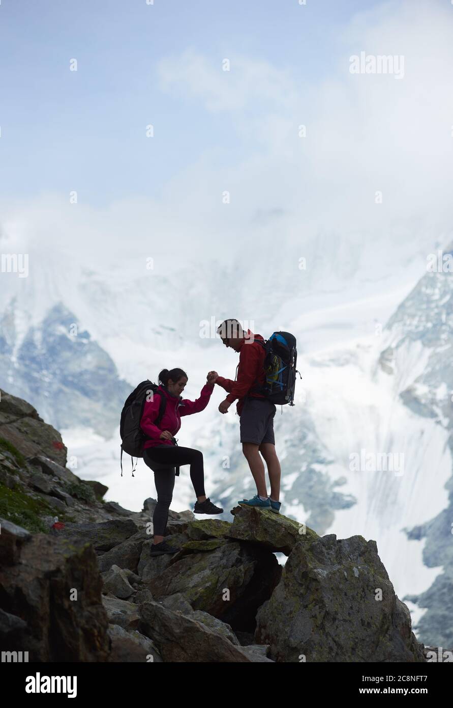 Wanderpaar Wandern in Schweizer Alpen, Mann hilft Frau zu klettern, Ober Gabelhorn in Wolken ist auf Hintergrund, Konzept des Abenteuers, Tourismus, Alpinismus in Alpen Stockfoto
