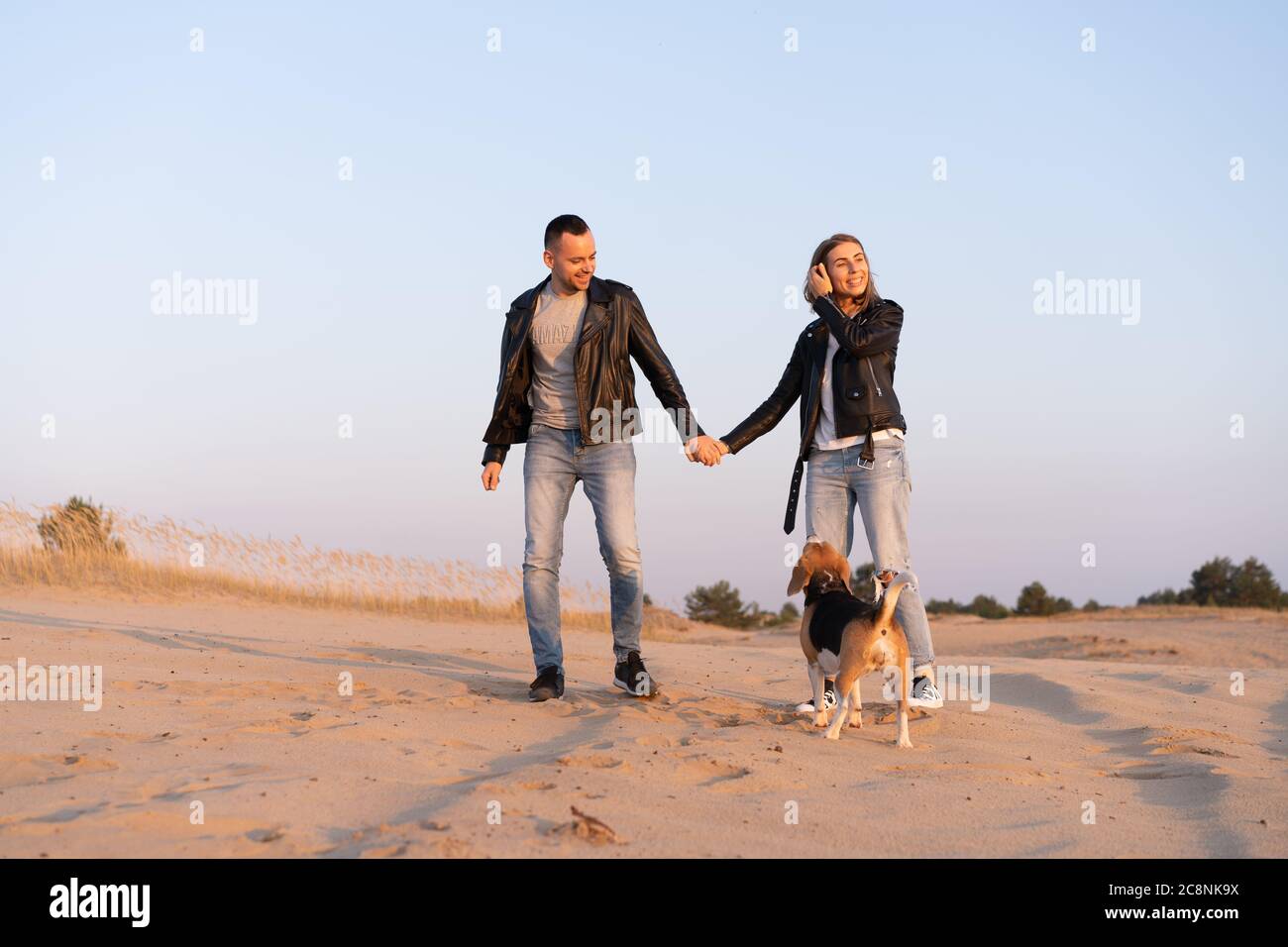 Junge schöne kaukasische Paar trägt Lederjacke und Jeans Spaziergänge Wüstensand mit Beagle Hund besten Freund. Familie mit Hund ohne Kinder in der Natur ruhen. Positive Emotionen glückliche Menschen Stockfoto