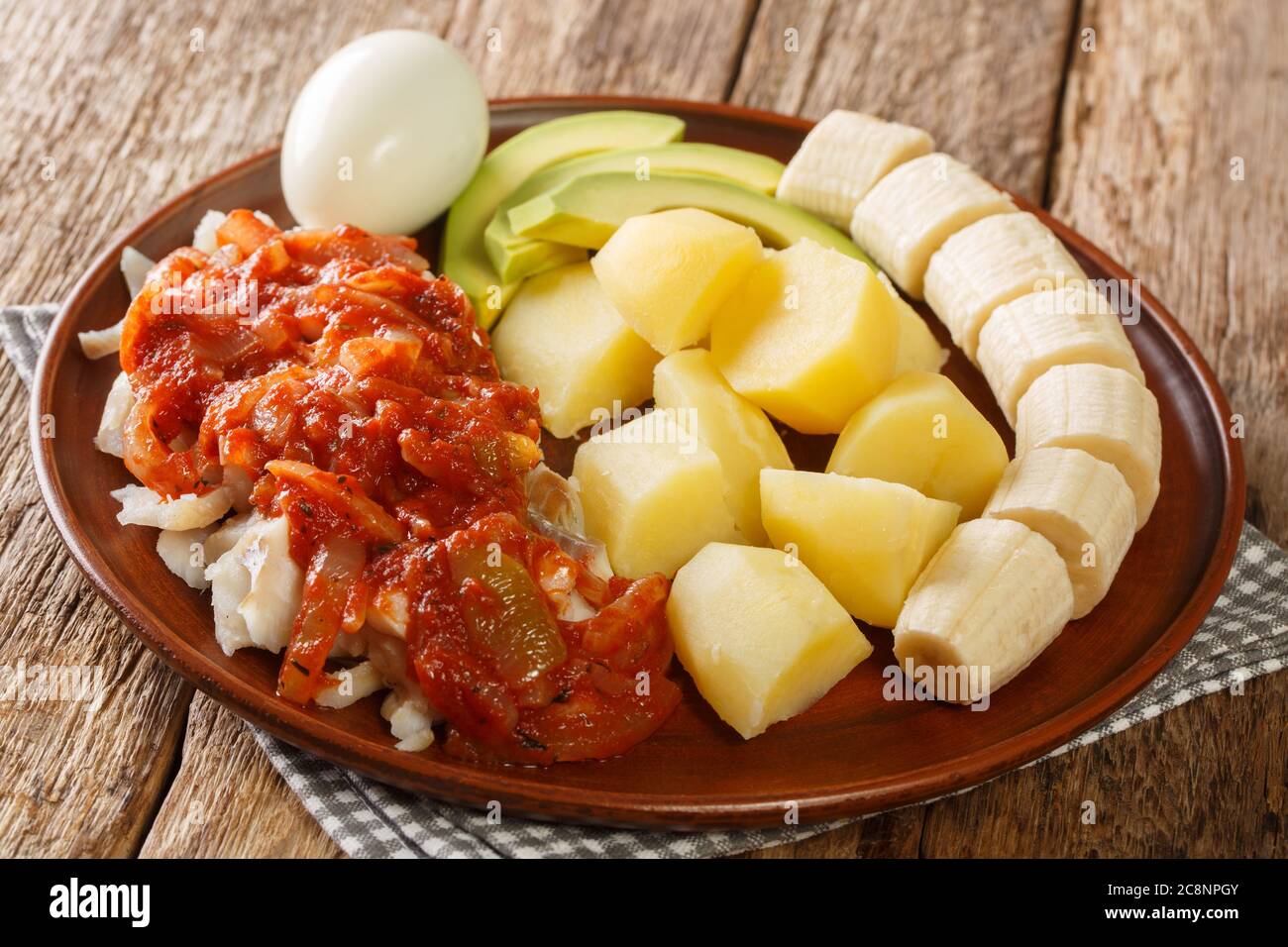 Gesundes Bermuda-Frühstück mit gesalzenem Kabeljau mit Zwiebeltomate-Sauce, gekochten Kartoffeln, Eiern, frischer Banane und Avocado in Nahaufnahme auf einem Teller auf dem Tisch. hor Stockfoto