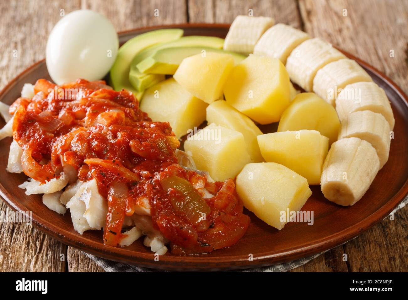 Köstliches Bermuda-Frühstück aus gesalzenem Kabeljau mit Zwiebeltomatensoße, gekochten Kartoffeln, Eiern, Banane und Avocado in Nahaufnahme auf einem Teller auf dem Tisch. Stockfoto