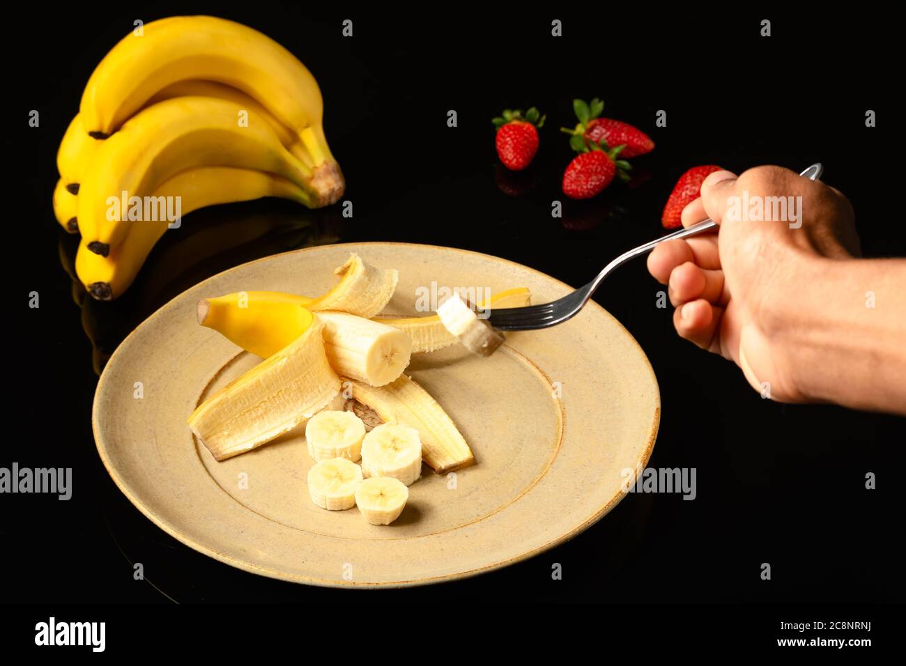 Banane auf einer Keramikplatte Bananenbündel am Boden und eine Hand mit einer Gabel Kommissionierung ein Stück Obst, in schwarzem Hintergrund fotografiert Stockfoto