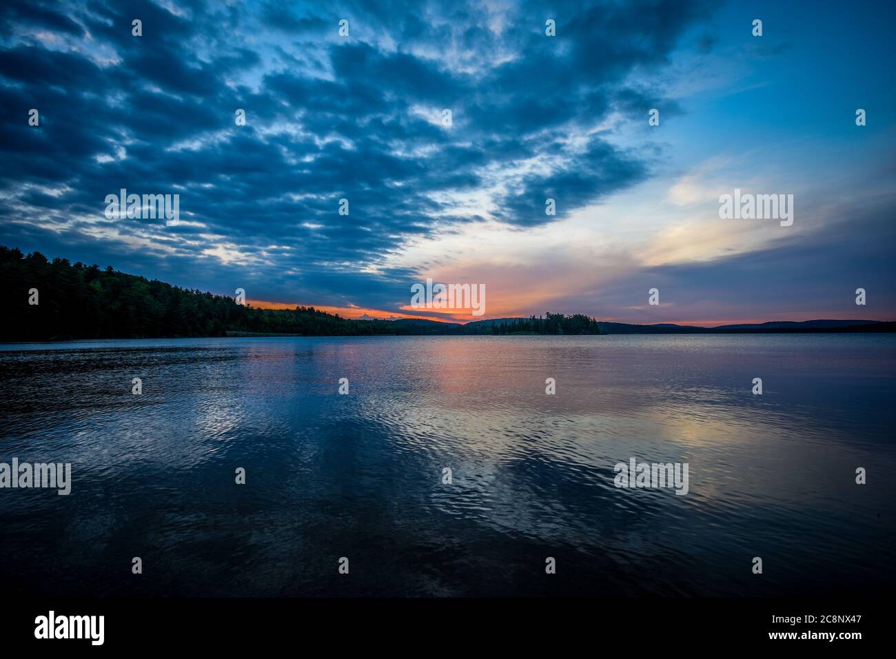 Sonnenaufgang, mit der Sonne durch den Baum in den Wald am fernen Ufer eines ruhigen Sees, die Reflexion eines bewölkten Himmels, an einem Sommermorgen. Stockfoto