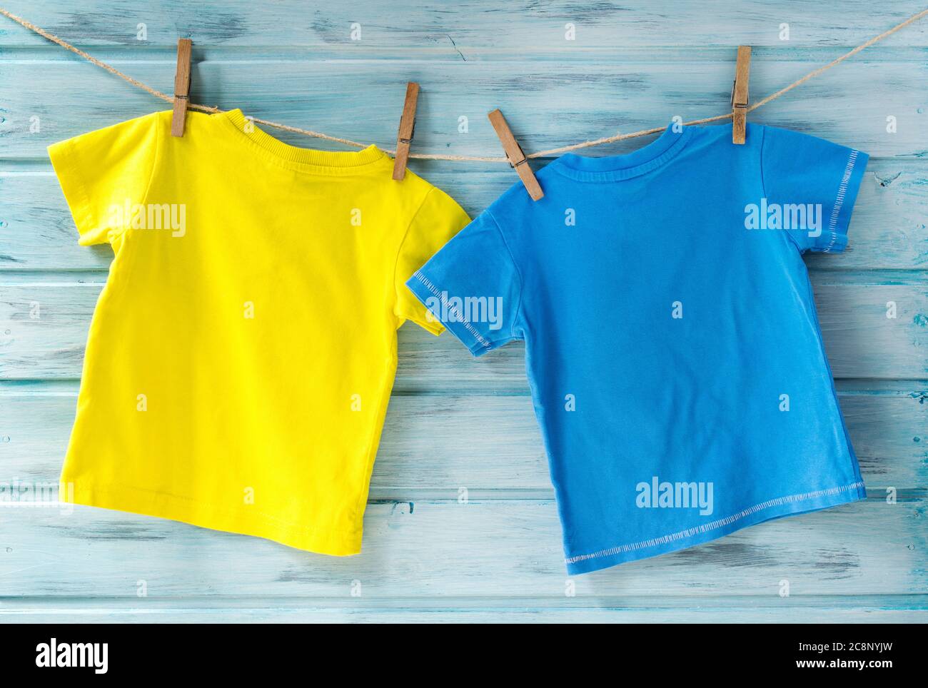 Zwei helle Baby-T-Shirts, die auf einer Wäscheleine auf einem blauen Holzhintergrund hängen Stockfoto