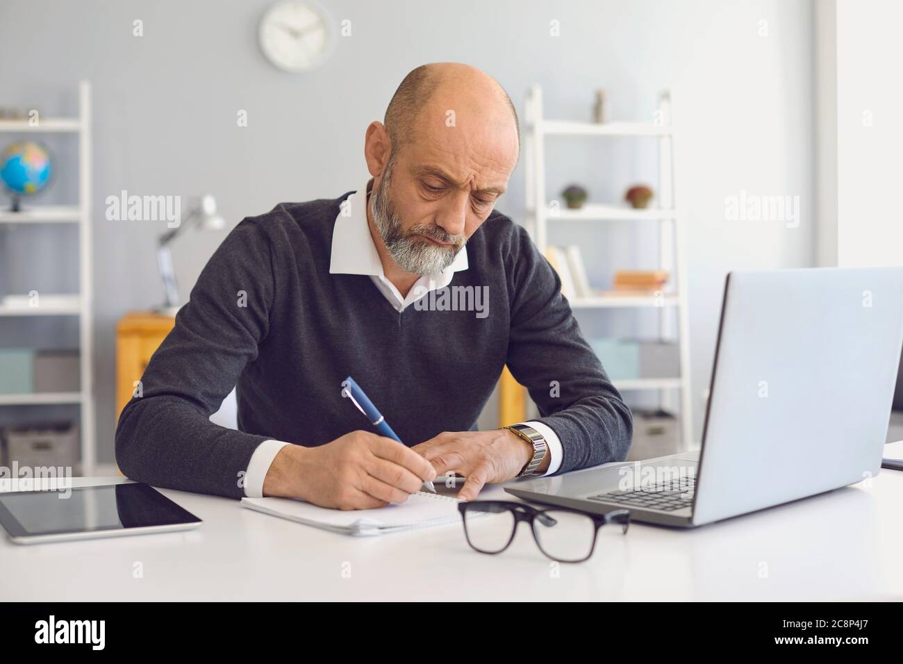 Arbeiten und Studieren von zu Hause aus. Durchdachter reifer Mann, der sich während eines Online-Meetings oder Webinars Notizen gemacht hat Stockfoto