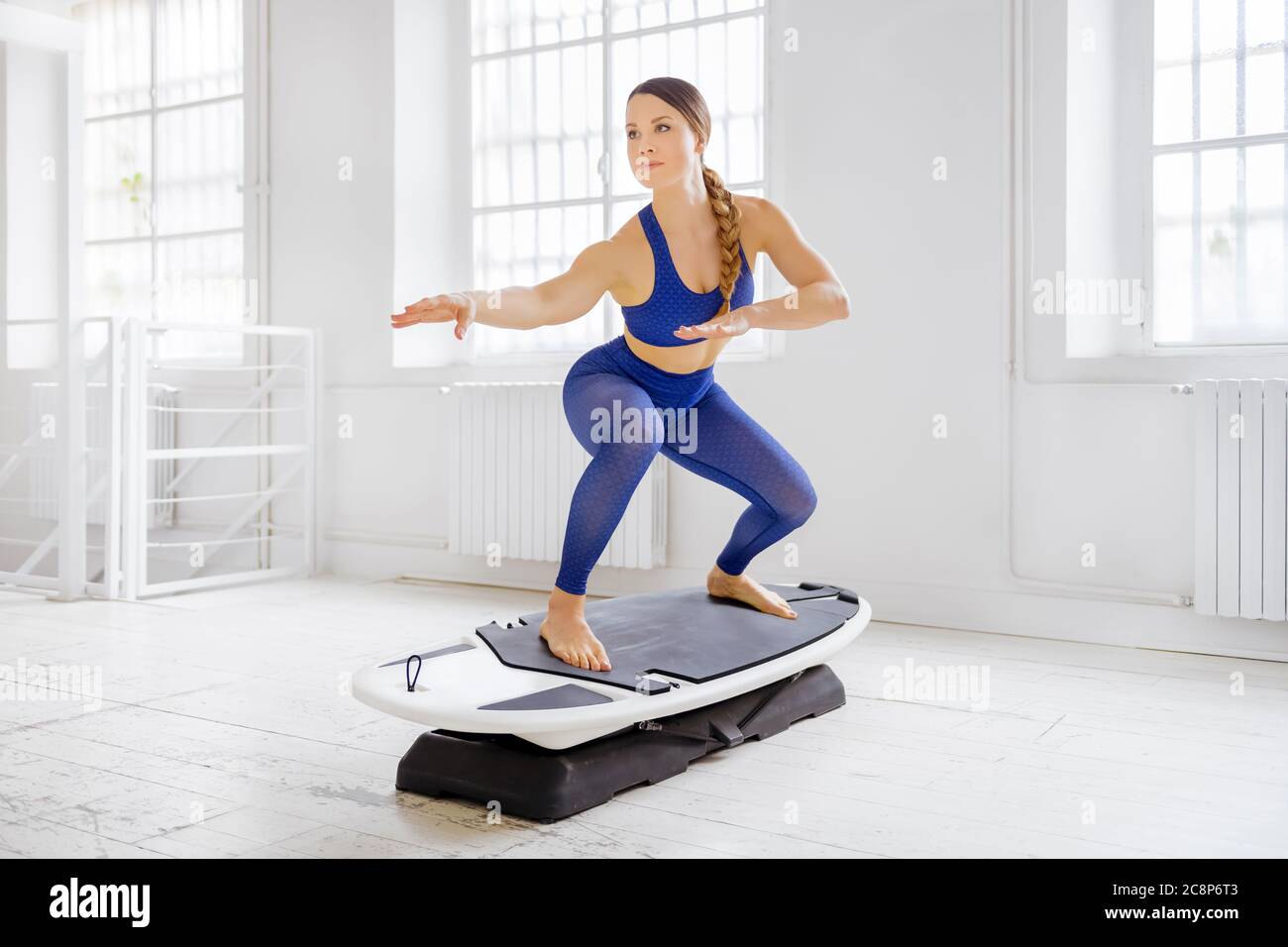 Junge Frau macht ein Surfset regelmäßige Surfer Pose während ihres Fitness-Workout in einem High-Key-Fitnessstudio mit Copyspace in einem gesunden Lifestyle-Konzept Stockfoto