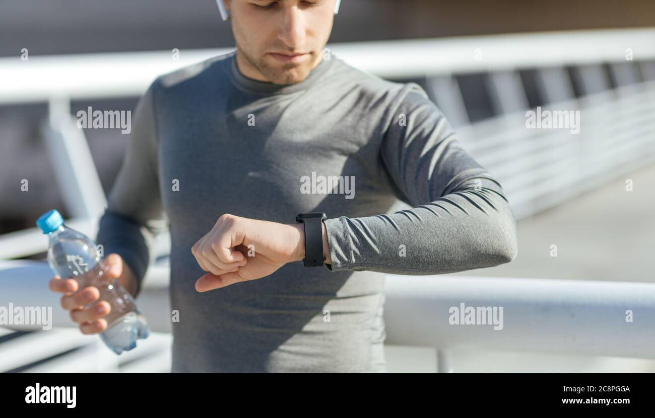 Junger Mann in kabellosen Kopfhörern mit Wasserflasche schaut auf Fitness Tracker im Stadion Stockfoto