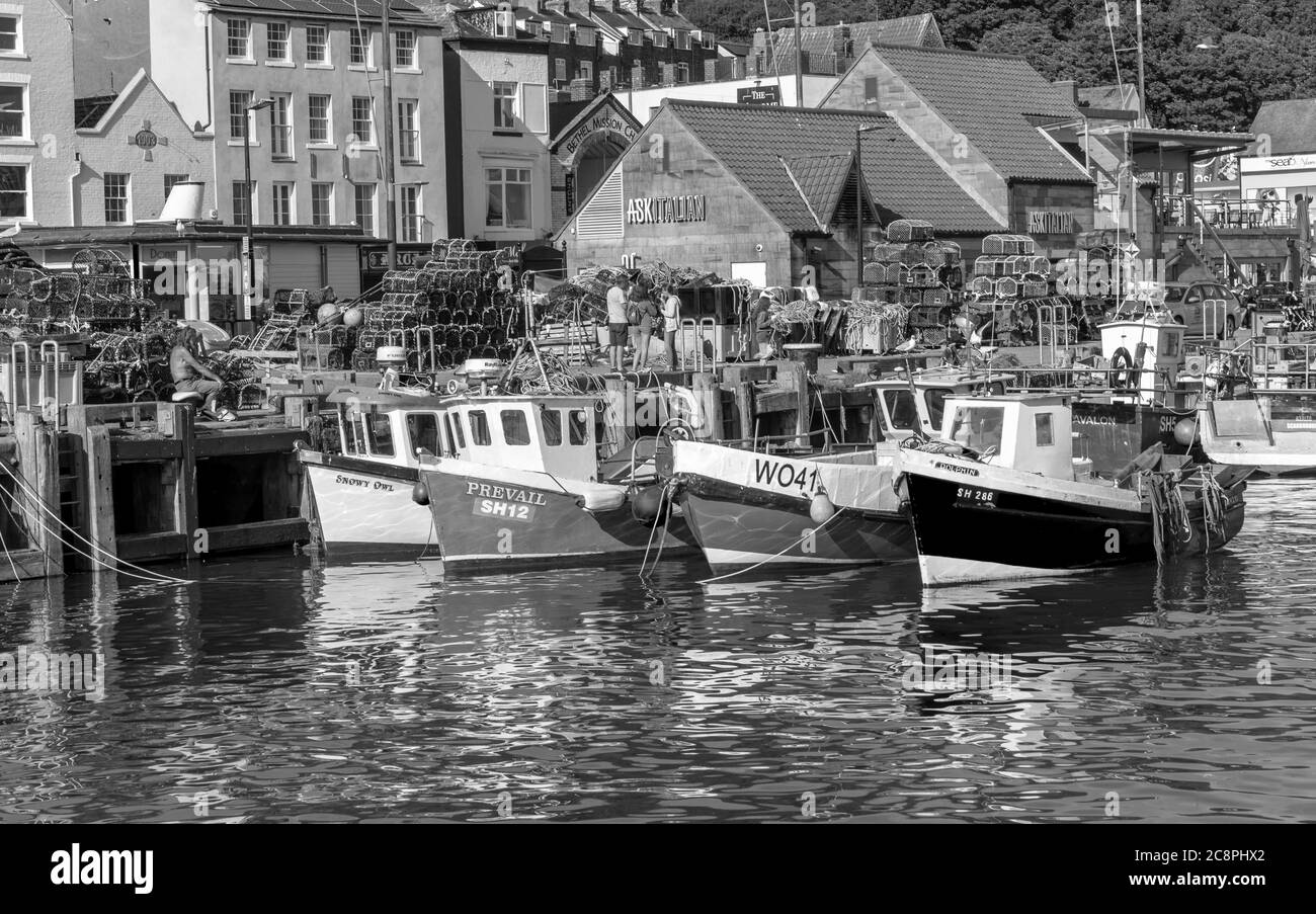 Fischerboote werden neben einem Kai vertäut, der mit Hummertöpfen gestapelt ist. Gebäude säumen die Uferpromenade und Touristen genießen die Umgebung. Stockfoto