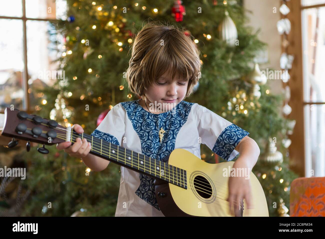 Lächelnder 4-jähriger Junge spielt Gitarre mit Weihnachtsbaum im Hintergrund Stockfoto