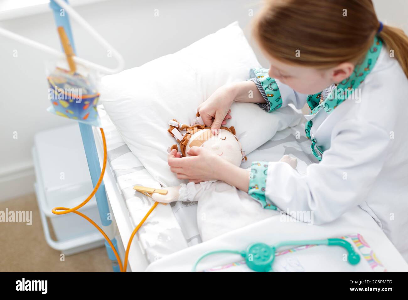 Junges Mädchen als Arzt gekleidet, das Gips auf den Kopf der Puppe in einem Krankenhausbett aus Make-believe setzt Stockfoto