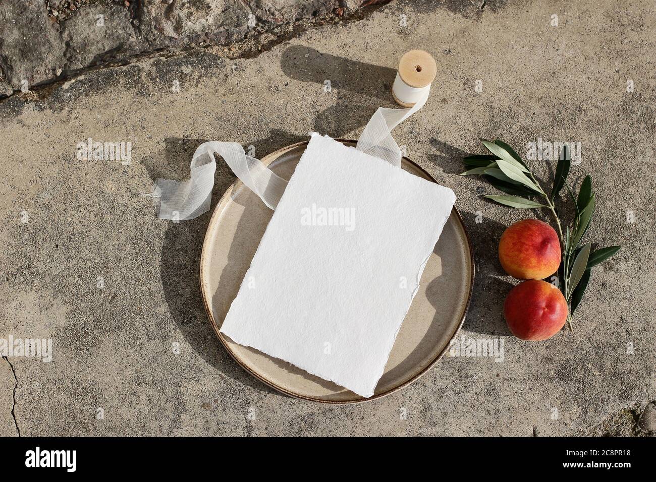 Sommer Hochzeit Schreibwaren. Unbeschriftete Grußkarte auf Keramikplatte im Sonnenlicht. Seidenband, Pfirsich Obst und Olivenzweig auf Beton Hintergrund Stockfoto