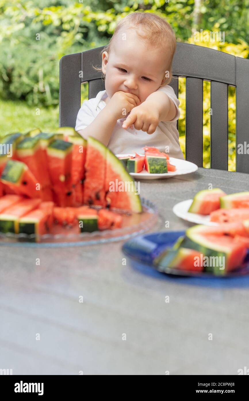 Niedlicher Junge isst und zeigt seine Hand auf eine Wassermelone, die auf dem Tisch liegt. Vertikal. Stockfoto