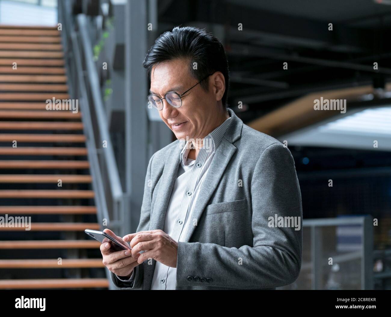 Chinesischer Geschäftsmann in einer Jacke, lächelnd und in seiner Hand auf das Handy schauend. Stockfoto