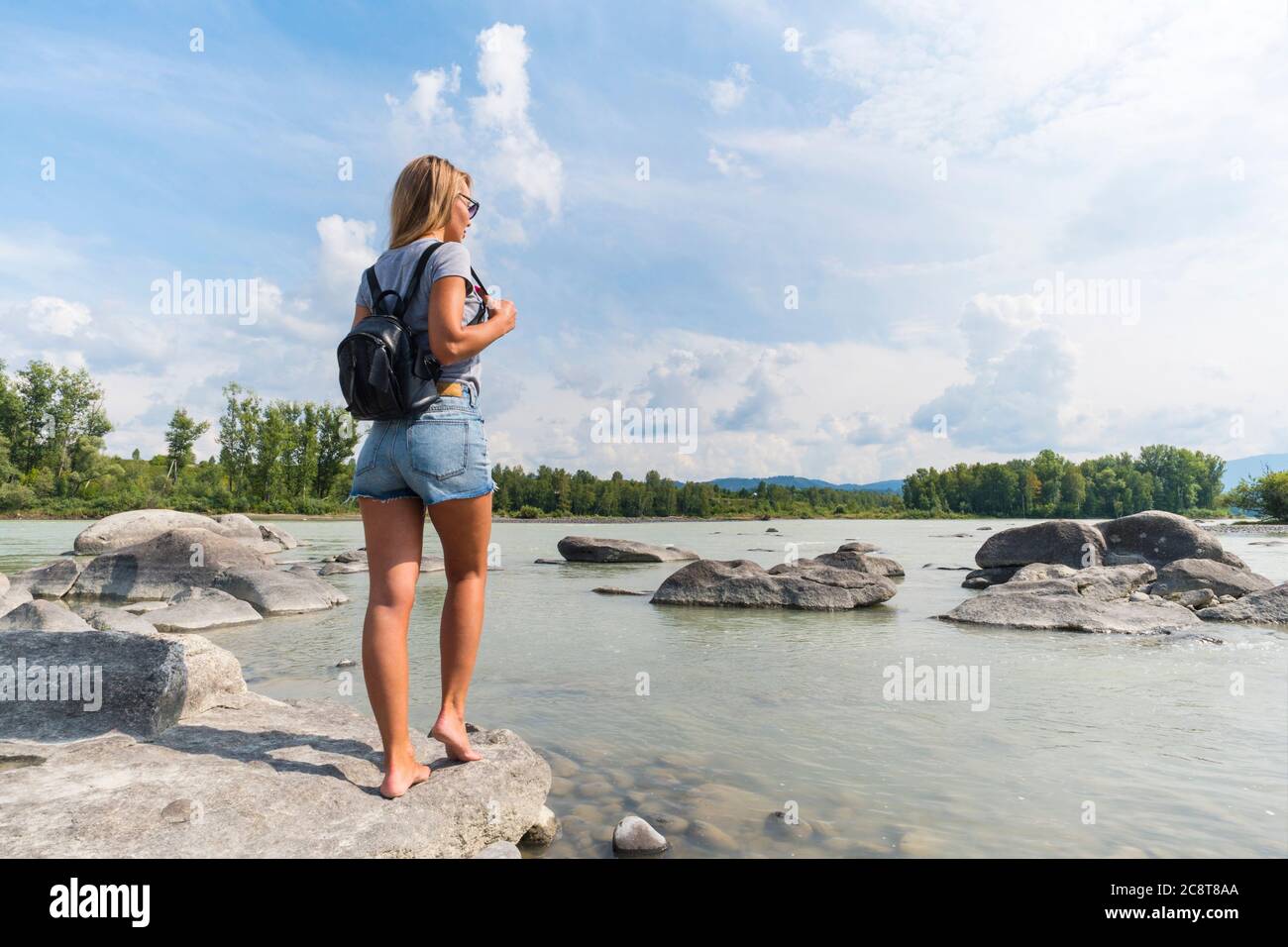 Blonde in schönen Denim-Shorts mit einem schwarzen Rucksack, der am Fluss auf den Felsen steht. Porträt von der Rückseite des Mädchen Reisenden. Fotoreporse Stockfoto