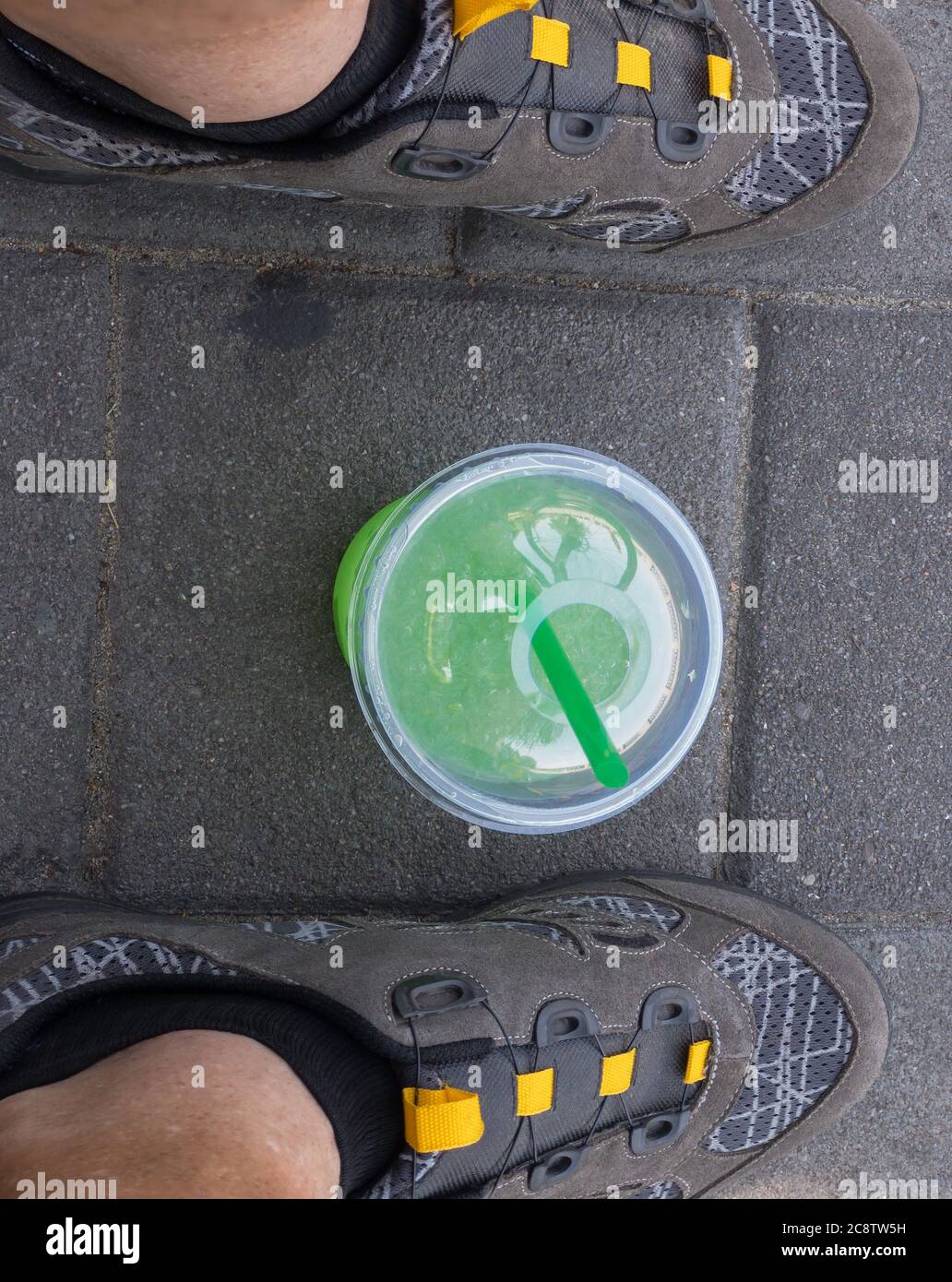 Draufsicht auf Plastikbecher mit grünlicher Flüssigkeit (Mojito) und Cocktail-Rohr innen, stehend auf Bürgersteig zwischen menschlichen Füßen Radschuhe Stockfoto