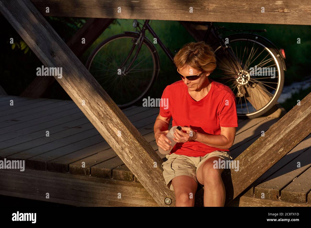 Eine Frau mittleren Alters sitzt auf einer Holzbrücke, ruht nach dem Radfahren und trinkt Wasser. Stockfoto