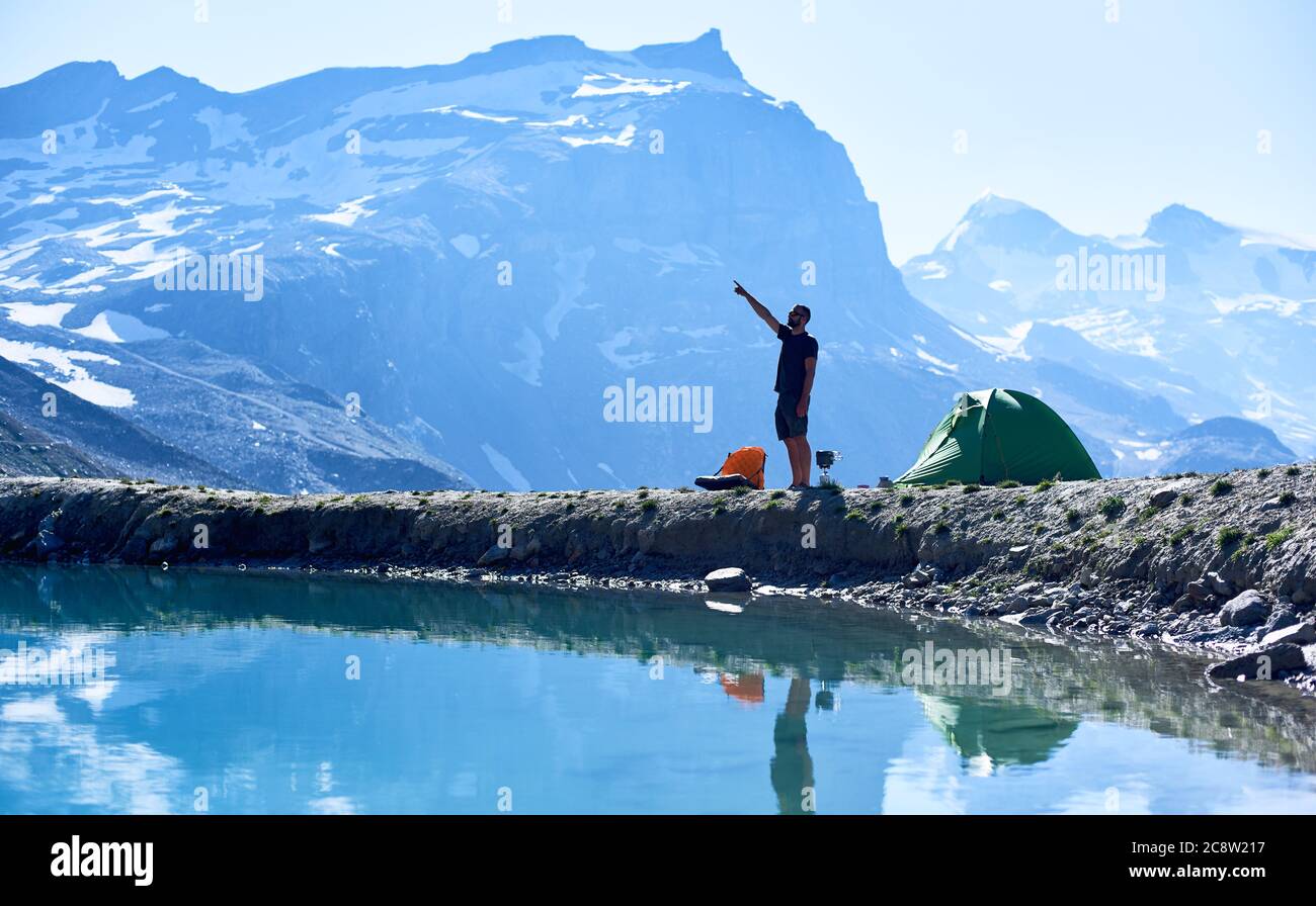 Seitenansicht des Reisenden in kurzen Hosen, die auf Berge zeigen, während sie am wunderschönen blauen See stehen. Tourist Zelt und junger Mann in kristallklarem Wasser reflektiert. Konzept von Reisen, Wandern und Camping. Stockfoto