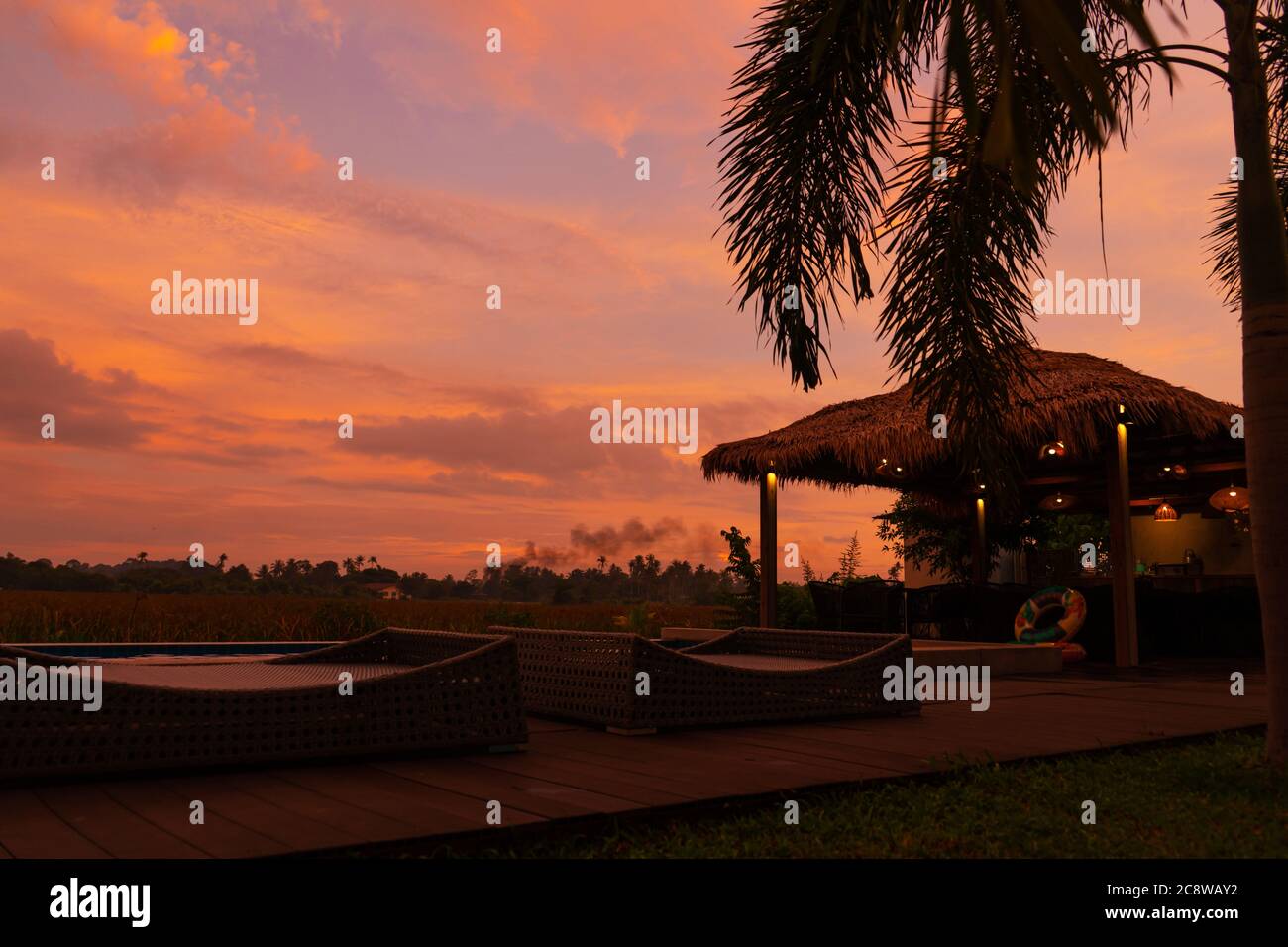 Schöner orange feuriger Sonnenuntergang in den Tropen. Straßendach von Heu, neben dem Pool und mit Blick auf das Reisfeld Stockfoto