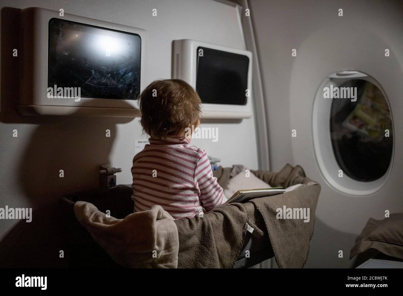 Konzept der komfortablen Flug mit Kleinkind. Beschäftigt Kleinkind sitzt in einem Liegekorb des Flugzeugs und ein Buch zu lesen Stockfoto