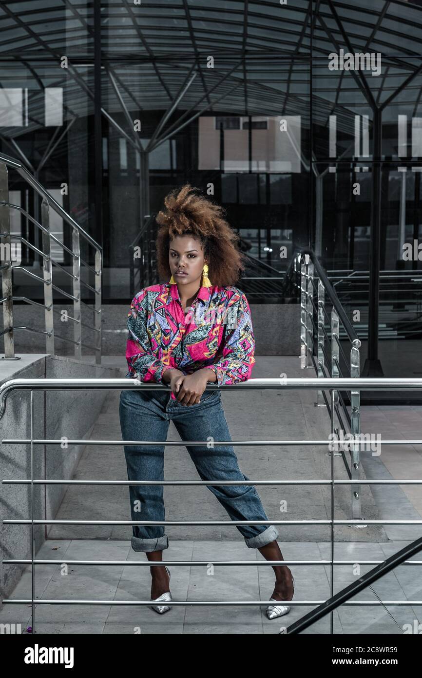 Ganzkörper junge afroamerikanische Frau mit afro Frisur tragen stilvolle helle Hemd mit Jeans auf Metall-Handlauf lehnt und Blick auf Kamera wi Stockfoto