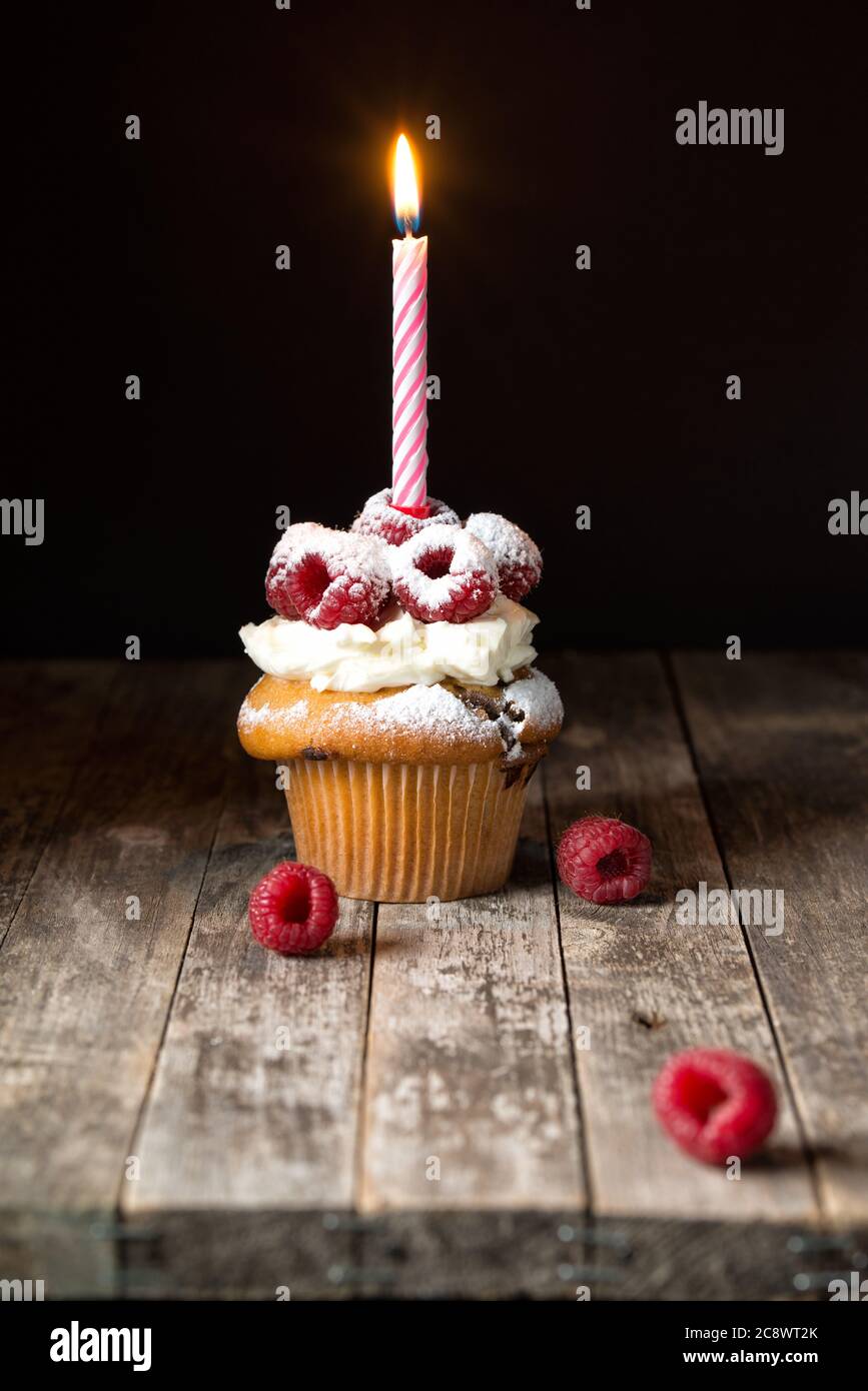 Muffin mit weicher Sahne und frischer Himbeere, gepudert mit Zucker und verziert mit brennender Kerze, Geburtstagskarte, rustikaler Hintergrund Stockfoto