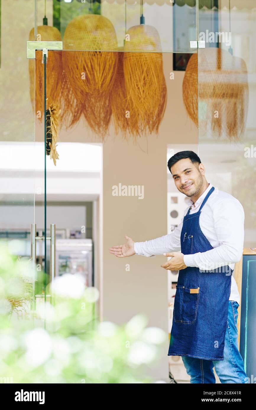 Positiver Cafe Kellner in Denim Schürze, der an Eingangstüren steht und Gäste einlädt Stockfoto