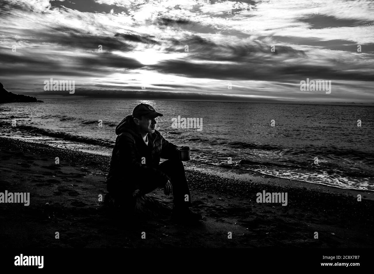 Morgen Tasse Tee. Mann sitzt auf einem Felsen und trinkt Tee bei Sonnenaufgang an einem Meeresstrand. Schwarz und Weiß, hoher Kontrast. Stockfoto