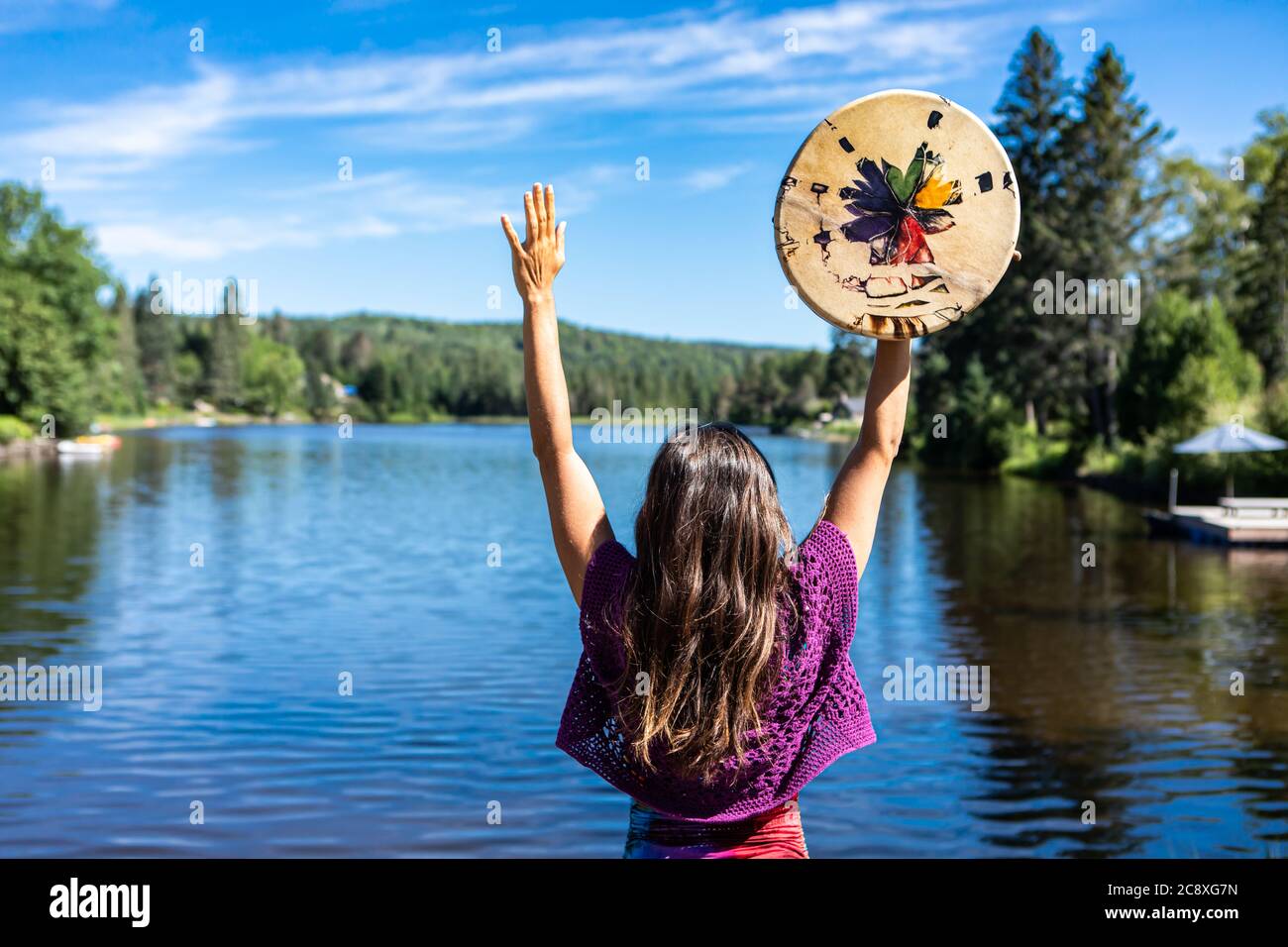 Junge Frau, die einen rituellen Tanz am Ufer eines kanadischen Sees aufführt und eine einheimische, handbemalte Trommel oder Tamburin in ihren erhobenen Händen hält. Stockfoto
