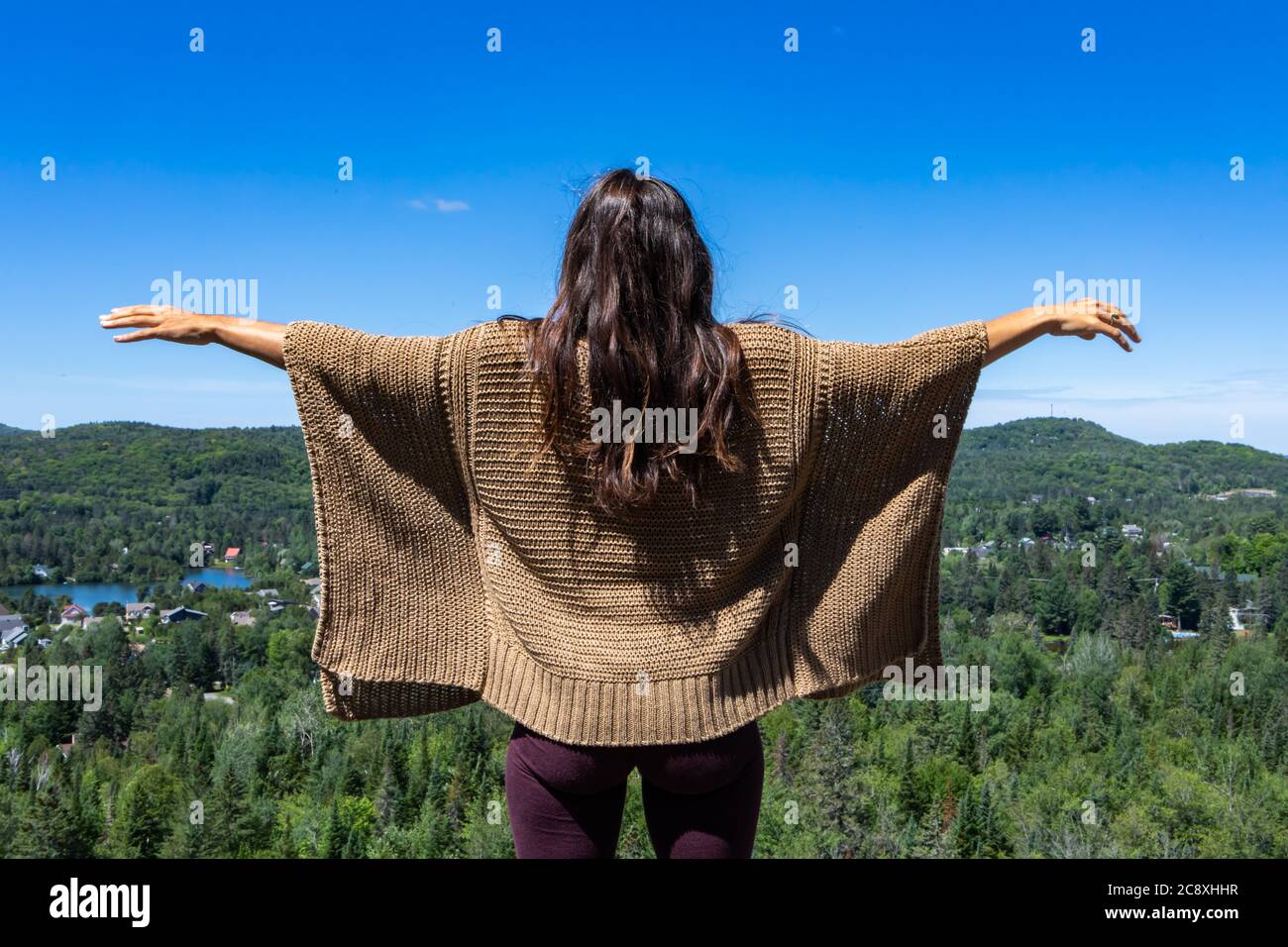 Langhaarige Frau in einem braunen Poncho-Pullover auf einem Hügel mit Blick auf einen kanadischen Wald, Hände oben in der Luft. Strahlend blauer Himmel. Stockfoto