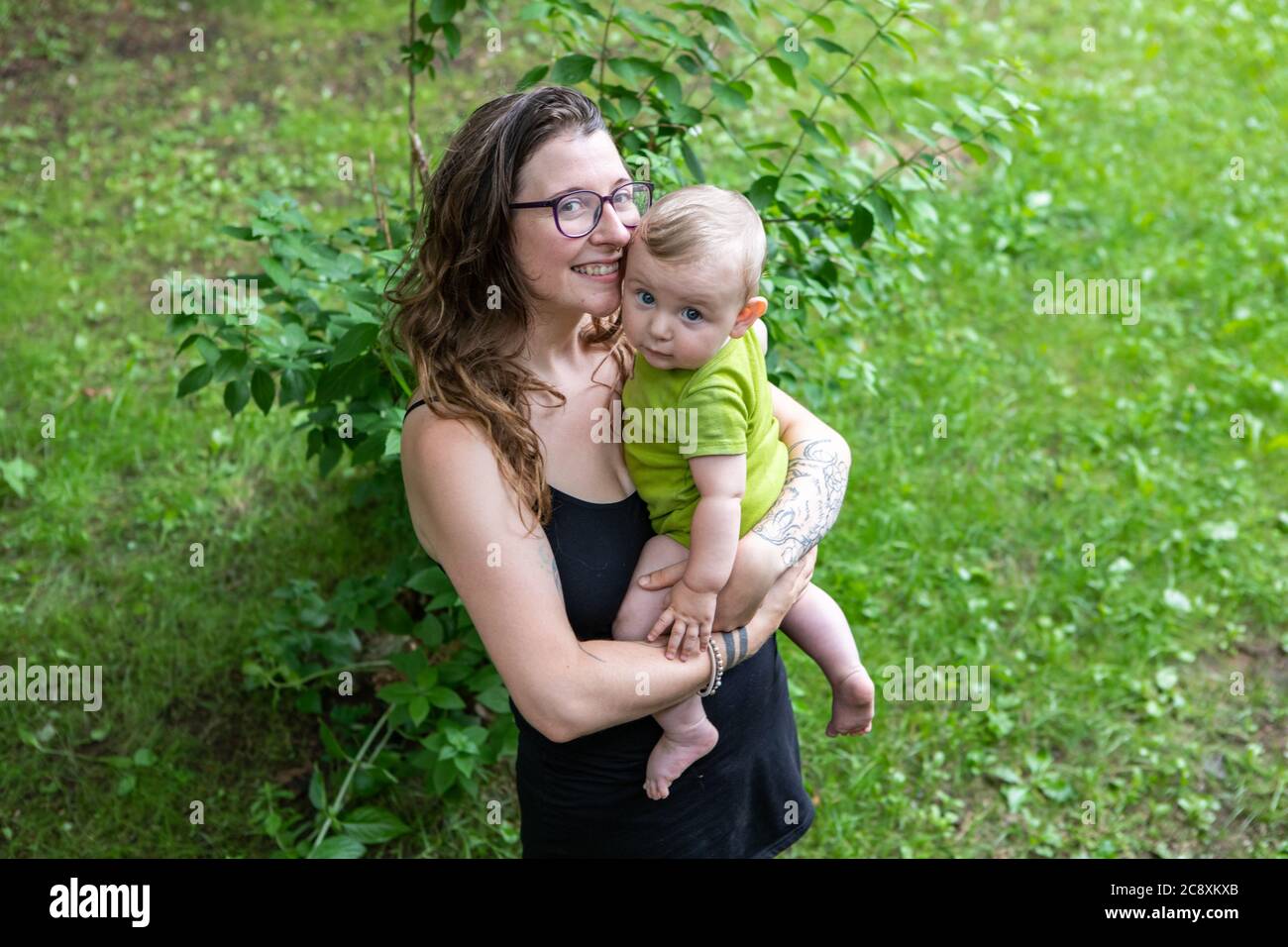 Porträt einer Mutter und ihres Babys, im Freien im Hinterhof. Mama hält ihren Sohn in den Armen, beide posieren für die Kamera, Mama mit einem großen Lächeln. Stockfoto