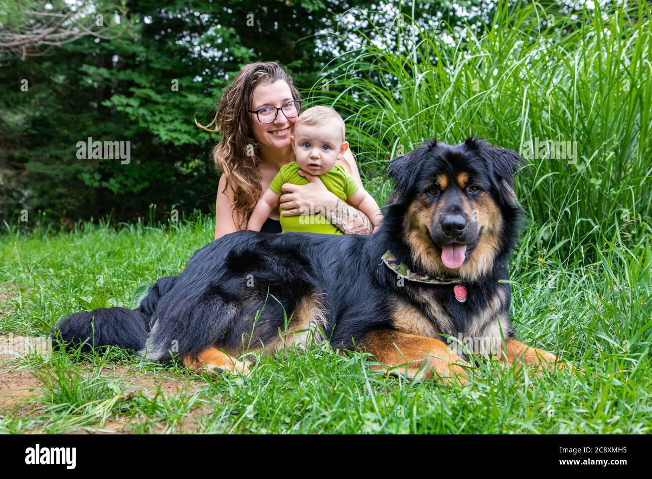 Mama, Baby-Sohn und große schwarze pelzigen Hund posiert für die Kamera im Outdoor-Porträt. Mama lächelt und hält ihr Baby, während sie alle in die Kamera schauen. Stockfoto
