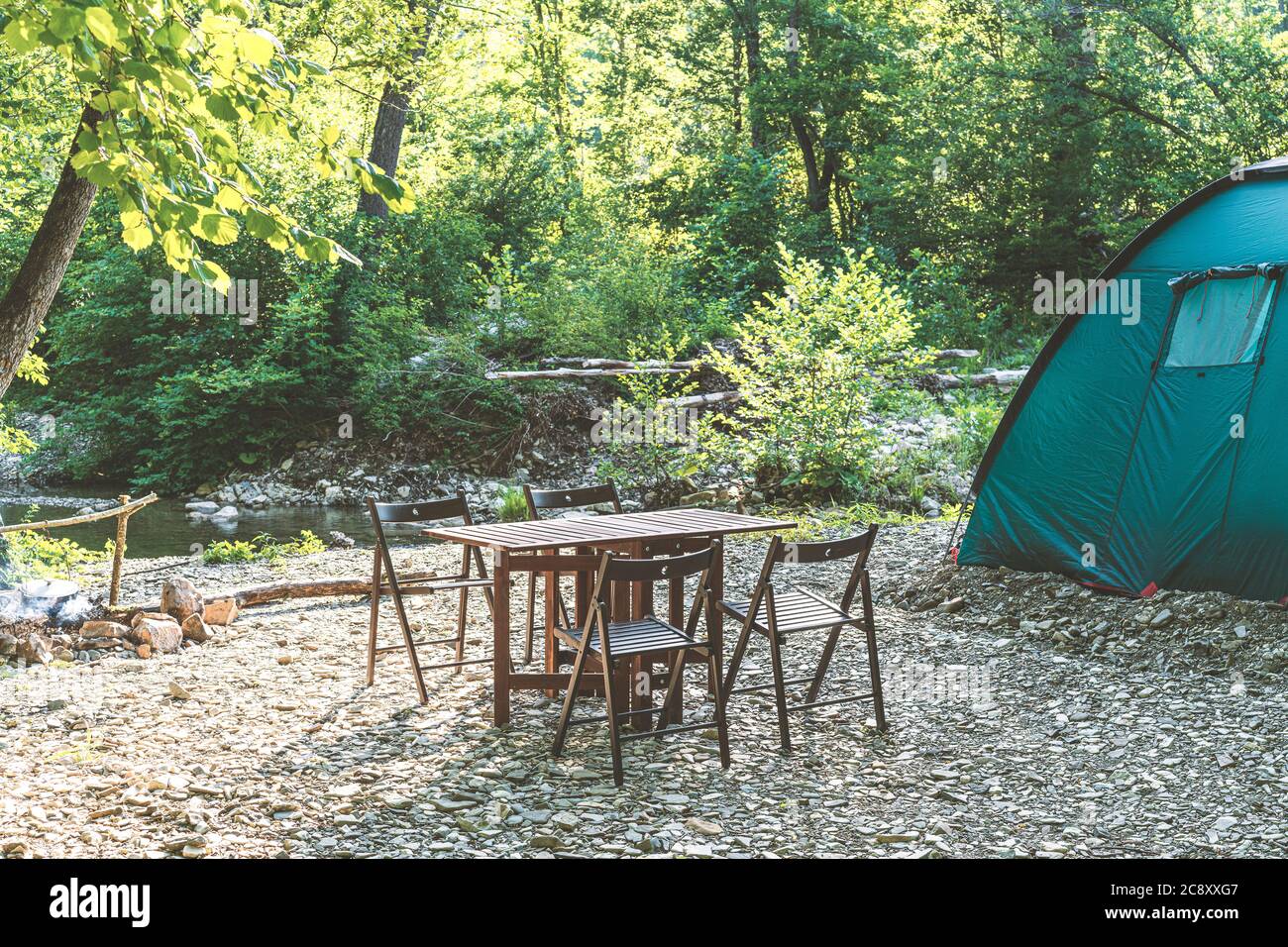 Camping am Strand des Bergflusses. Blaues Zelt, Lagertisch und Stühle im Wald. Sommerreisethema. Ruhestätte in der Natur. Stockfoto